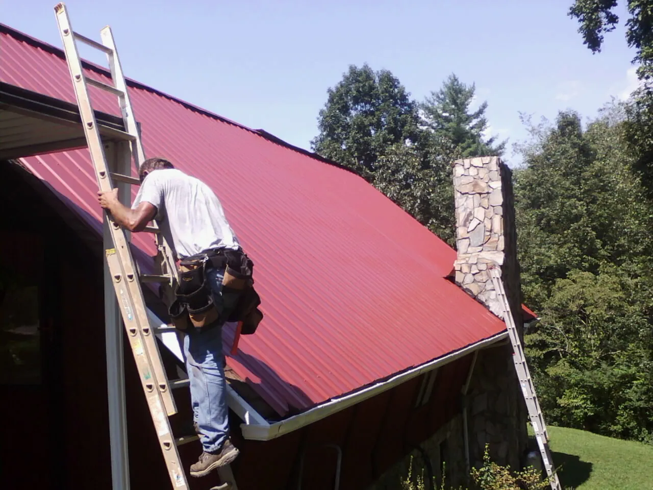 Roofer on a ladder beside a red metal roof and stone chimney — Soffit & Fascia Repair by Roofing Brothers in Stony Brook